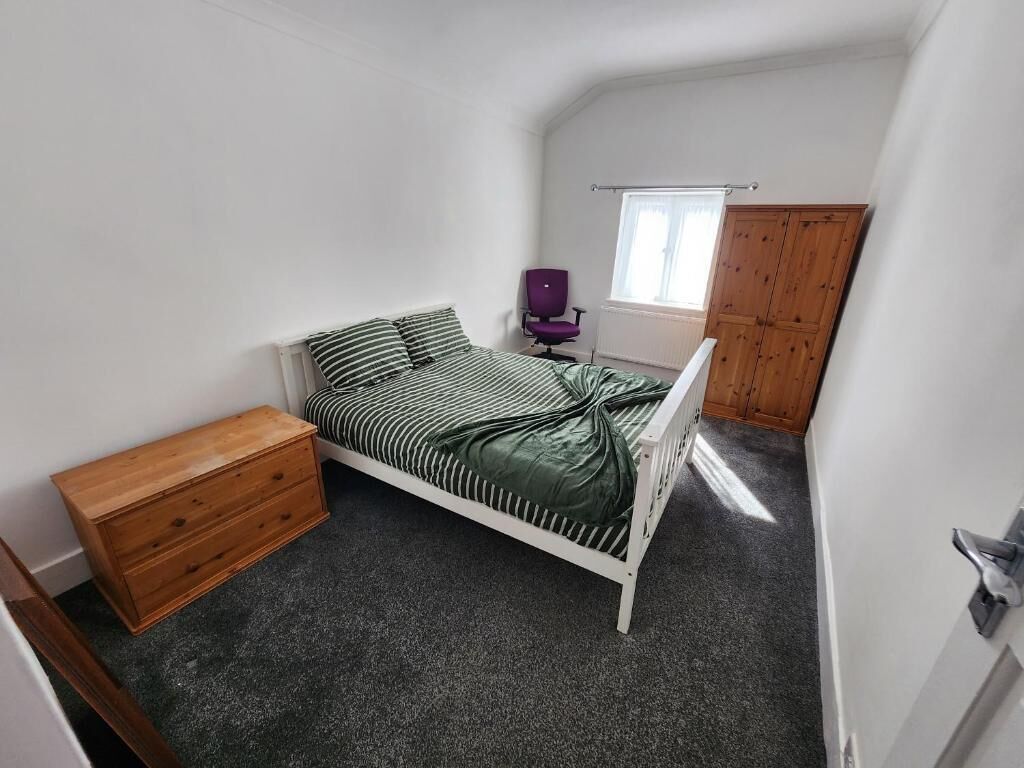Ein kleines Zimmer mit einem Einzelbett, einem Holzschrank, einem violetten Sessel und einem Fenster mit Blick auf ein Gebäude.