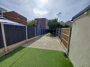 A paved backyard with artificial grass, a wooden fence, and a building in the background.