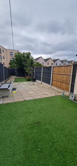 A residential backyard with a paved patio, a green lawn, and a wooden fence. - The Maple (Grays)