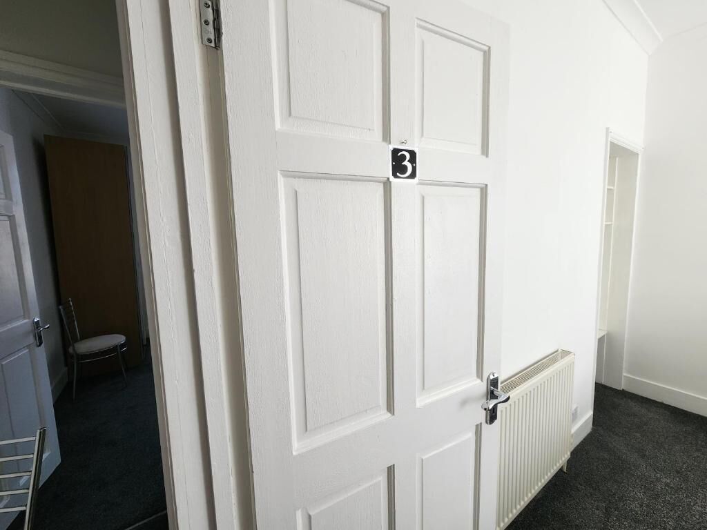 Una puerta blanca con una placa de número, un radiador y una silla parcialmente visible al fondo.