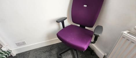 A purple office chair with wheels in a corner of a room with a white radiator and a grey carpet.