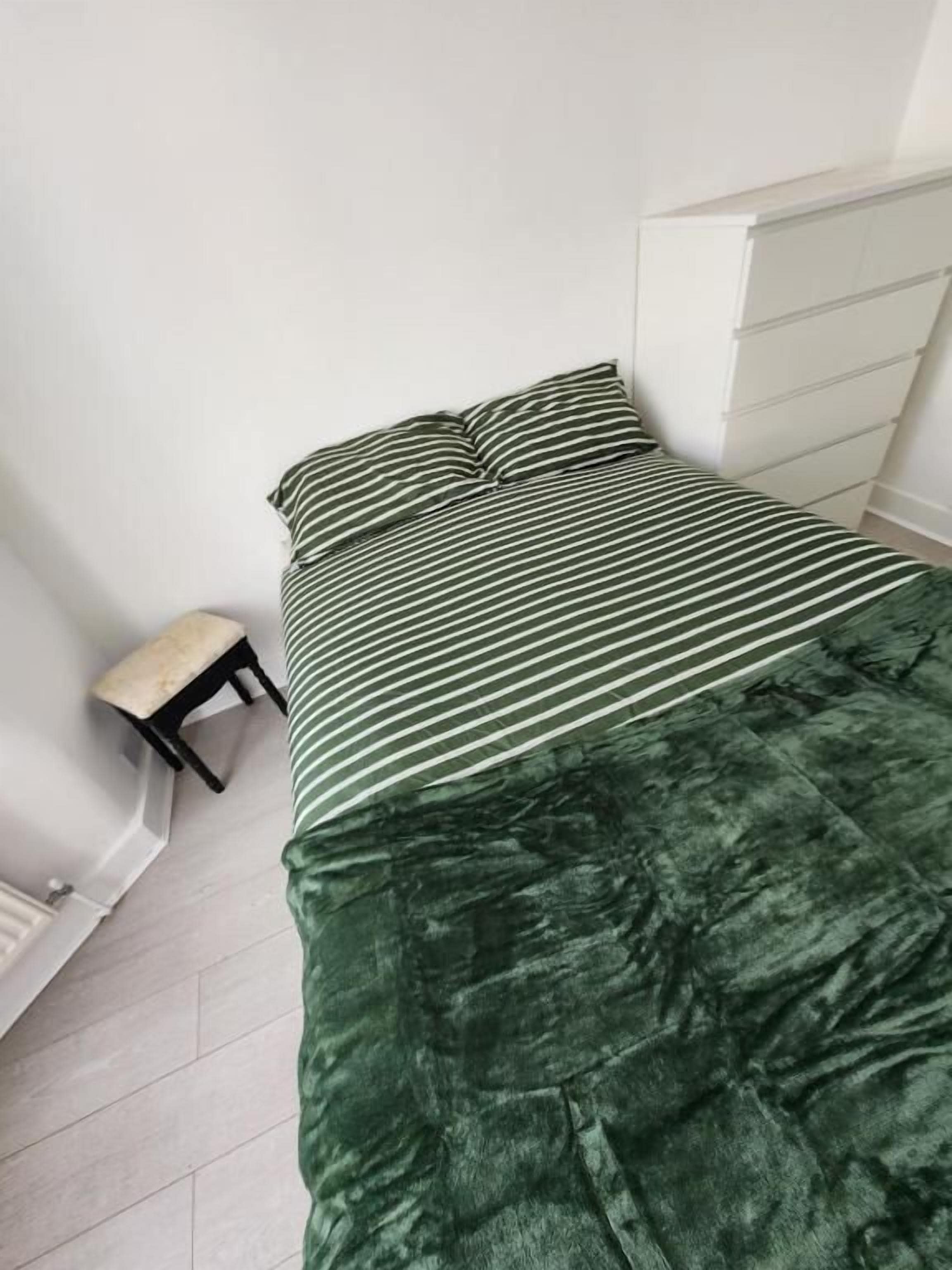 A bed with a striped bedspread and pillows, a small bench, and a white dresser in a room with white walls and a tiled floor.