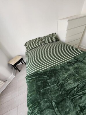 A bed with a striped bedspread and pillows, a small bench, and a white dresser in a room with white walls and a tiled floor.