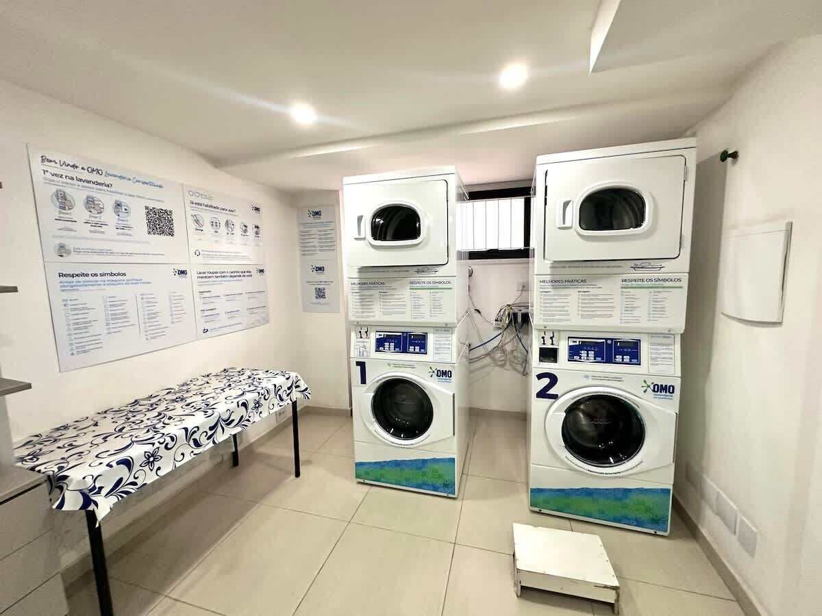 A laundry room with two stacked washing machines, a table with a patterned tablecloth, and informational posters on the walls.