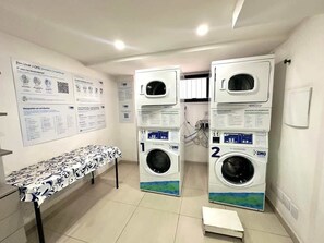 A laundry room with two stacked washing machines, a table with a patterned tablecloth, and informational posters on the walls. - Fortune Residence (São Paulo)