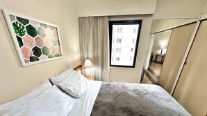 Una habitación de hotel con cama, ventana, lámpara y espejo.