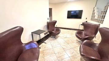 Uma área de espera com quatro poltronas de couro marrom, uma mesinha e uma televisão de tela plana na parede.