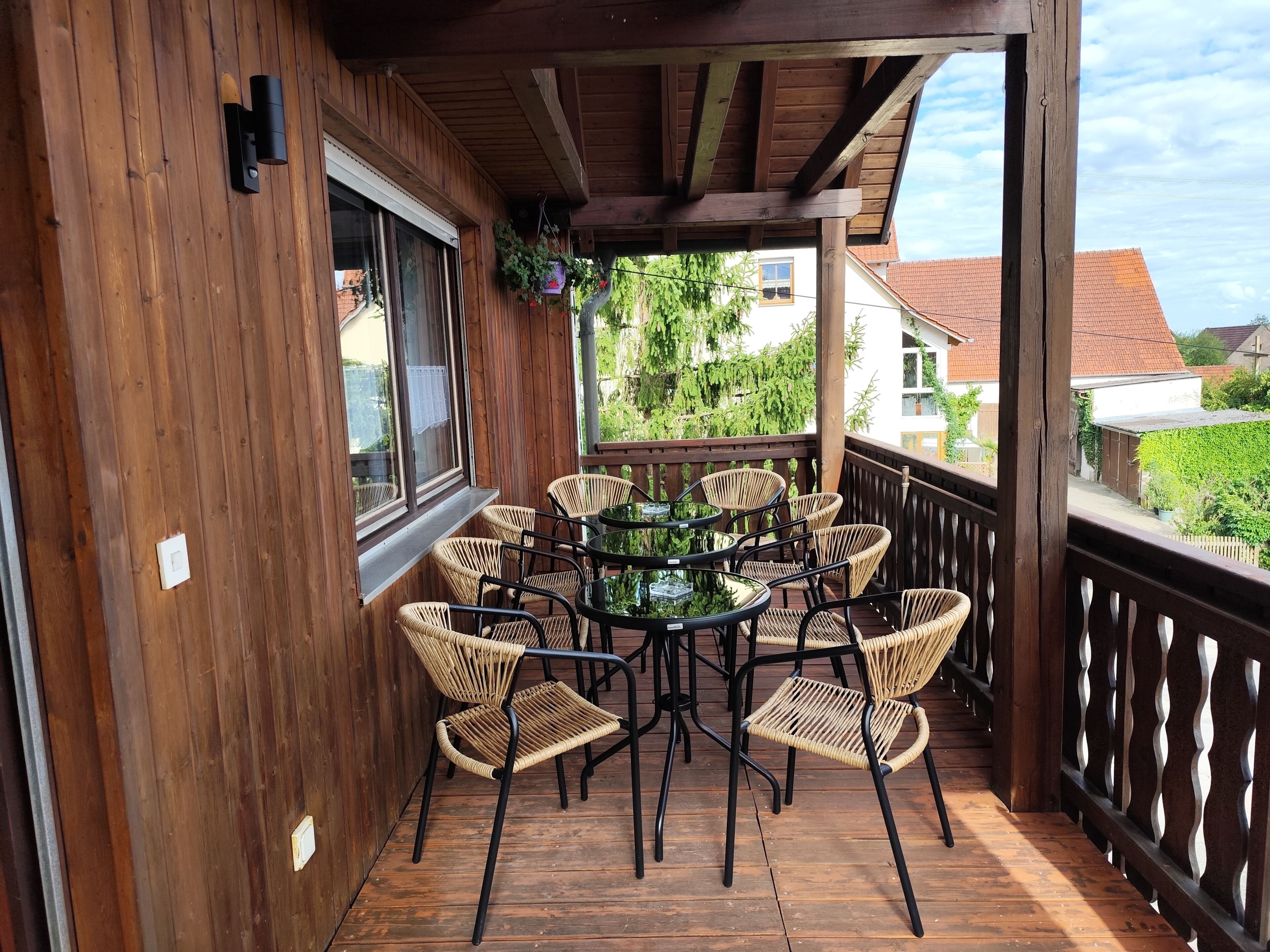 Une terrasse en bois avec une table ronde et des chaises en osier, une fenêtre avec un rideau et, au loin, une vue sur des maisons.