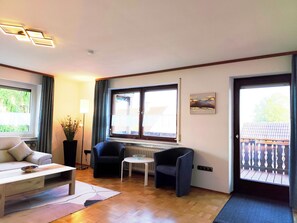 Ein Zimmer mit einer Couch, zwei Sesseln, einem kleinen Tisch und einem Heizkörper. Ein Fenster mit Vorhängen, ein Bild an der Wand und eine Tür mit Blick auf einen Balkon.