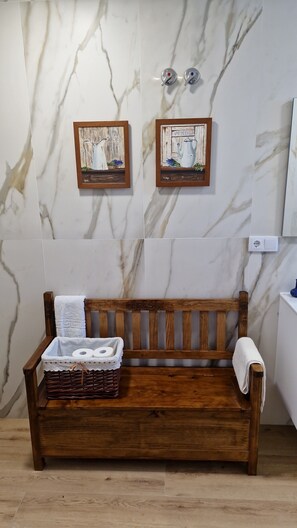 Eine hölzerne Sitzbank mit einem weißen Handtuch und einem Korbwäscheschrank in einem Badezimmer mit Marmorwänden und zwei gerahmten Gemälden.