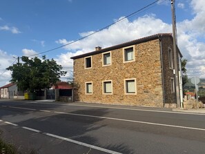 Un edificio de piedra de dos pisos con techo plano y una chimenea visible, con una calle en frente.