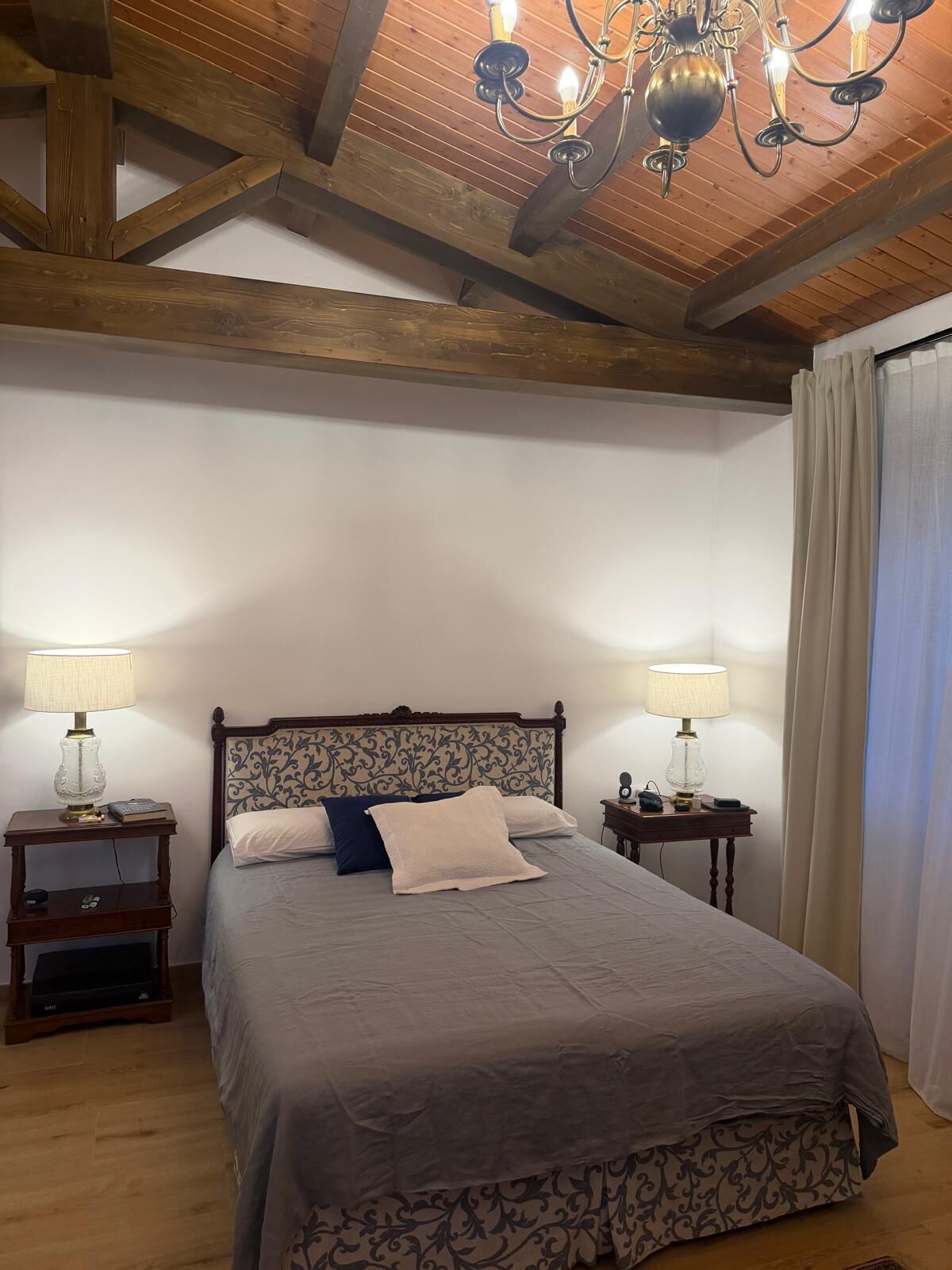 Un dormitorio con techo de madera, una cama con cabecera estampada, dos mesitas de noche con lámparas y una ventana con cortinas.