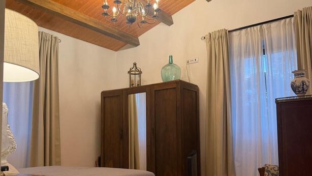 Ein Schlafzimmer mit Holzboden, einem Bett, einem Nachttisch, einer Kommode, einem Sessel und einem Fenster mit Vorhängen.
