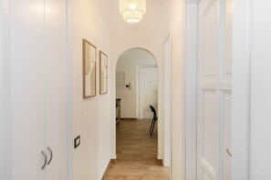 A hallway with white walls, a wooden floor, and a door leading to a room. There are framed pictures on the wall and a light fixture above. - La Maison Mikonos - superior apartment near the sea (Paola)