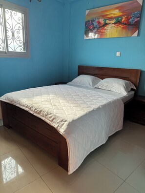 Une chambre à coucher avec un lit en bois, du linge de lit blanc, une fenêtre ornée de barreaux en fer décoratifs et un tableau coloré accroché au mur.