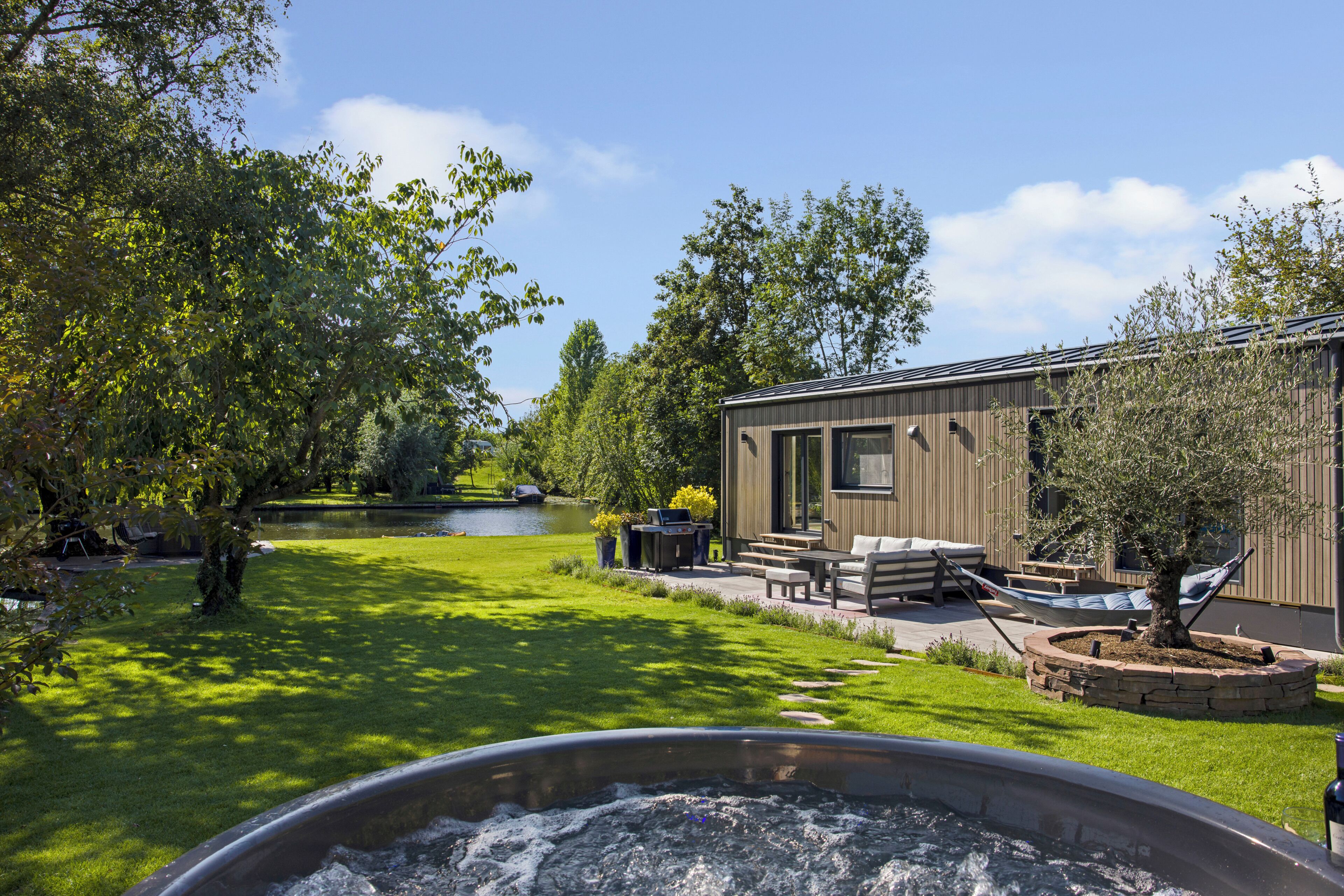 Een hottub in de voorgrond, een gazon, een vijver en een houten gebouw met terras.