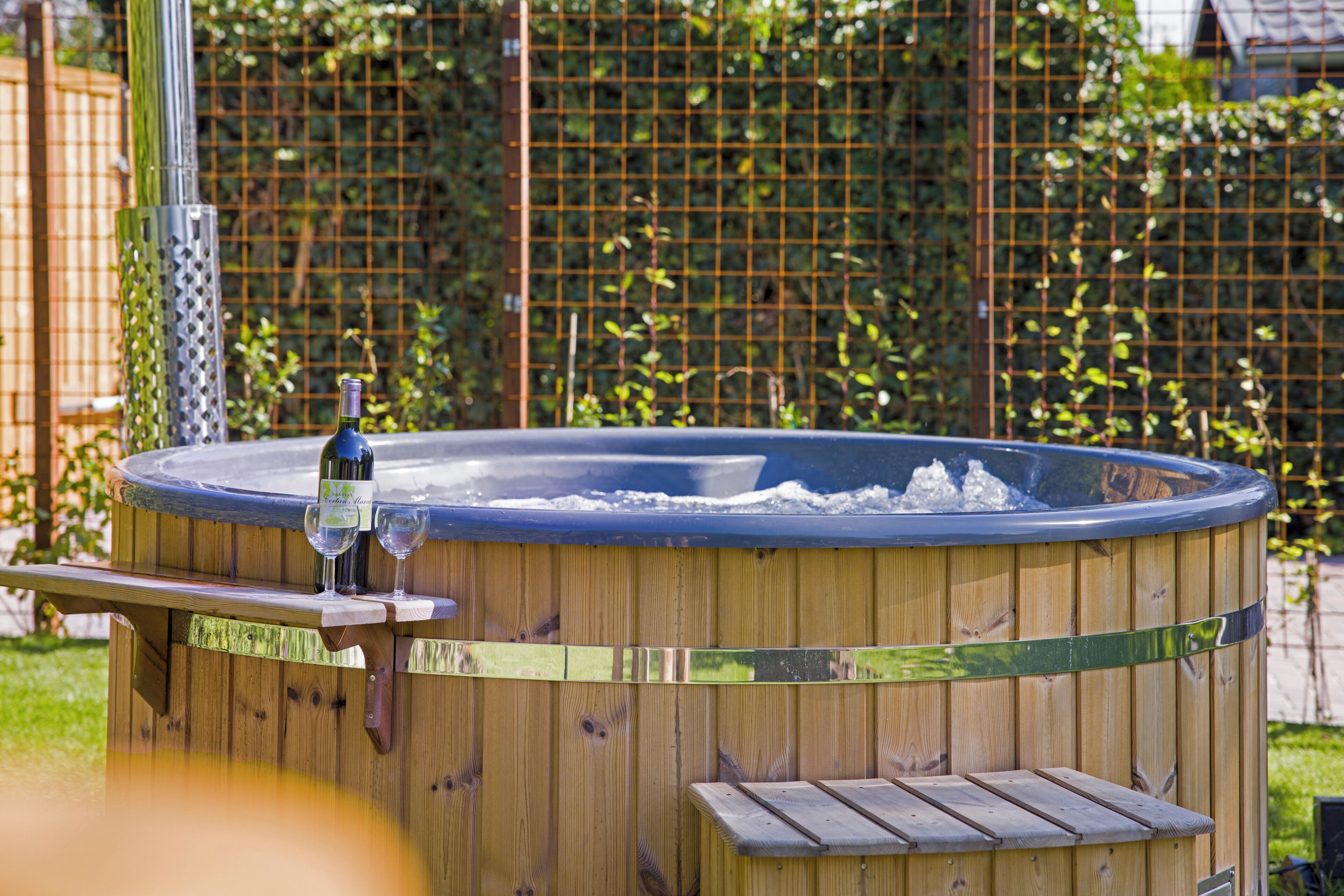 A wooden hot tub with a metal frame, two empty glasses, and a bottle of wine.