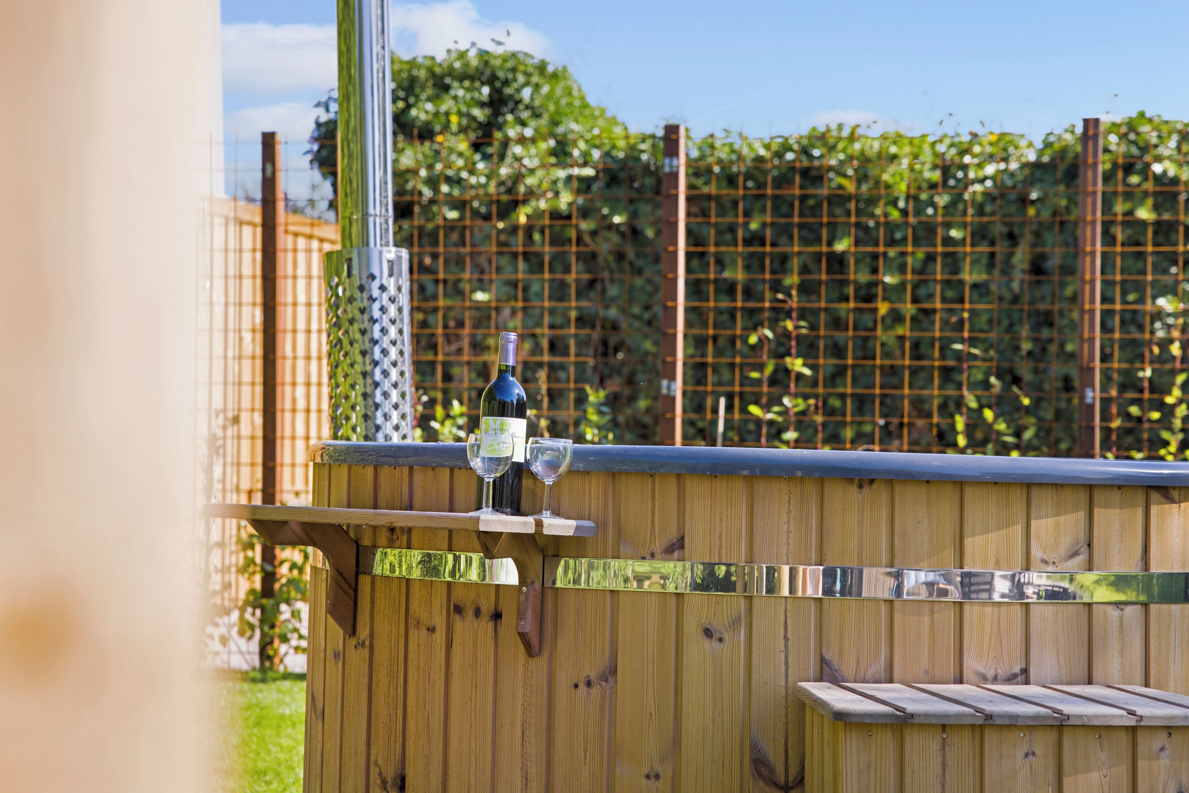 A wooden outdoor seating area with a bottle of wine and two glasses on a table.