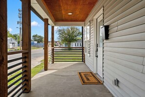 A covered porch with a wooden railing, a welcome mat, and a view of the street and houses in the background. - Hot Tub + Fire Pit Yard · Near Fountain Square (Indianapolis)