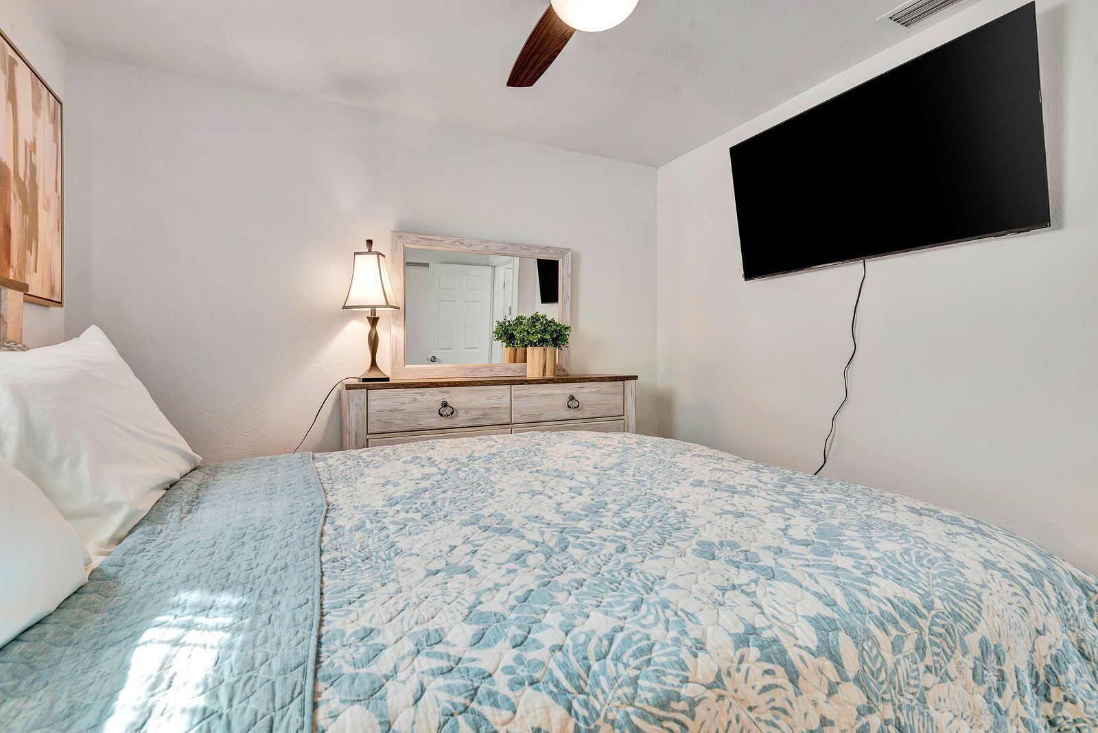 Een slaapkamer met een bed, een dressoir, een spiegel, een aan de muur bevestigde televisie en een plafondventilator.