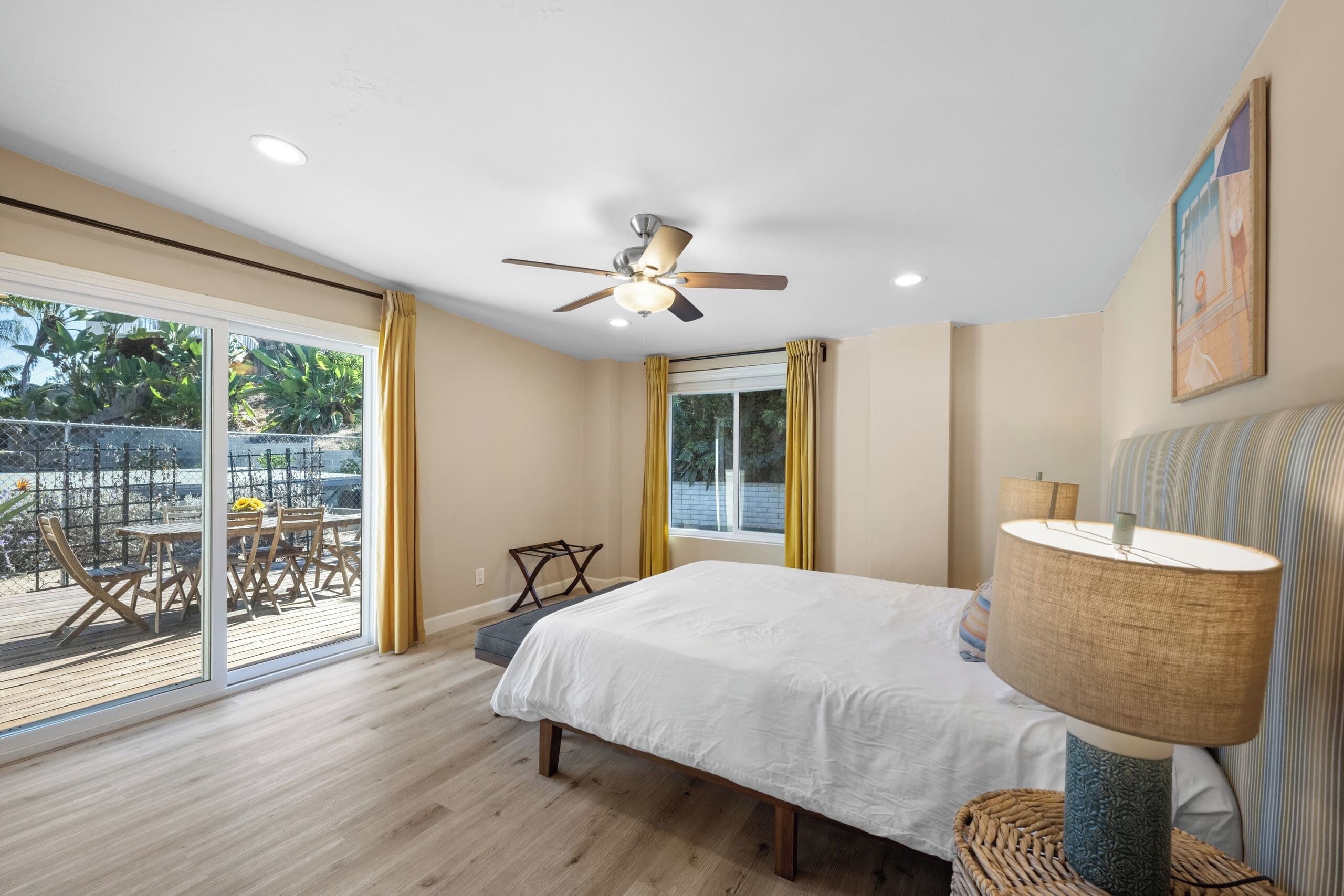 Quarto com cama, ventilador de teto, janela com cortinas e vista para um terraço.