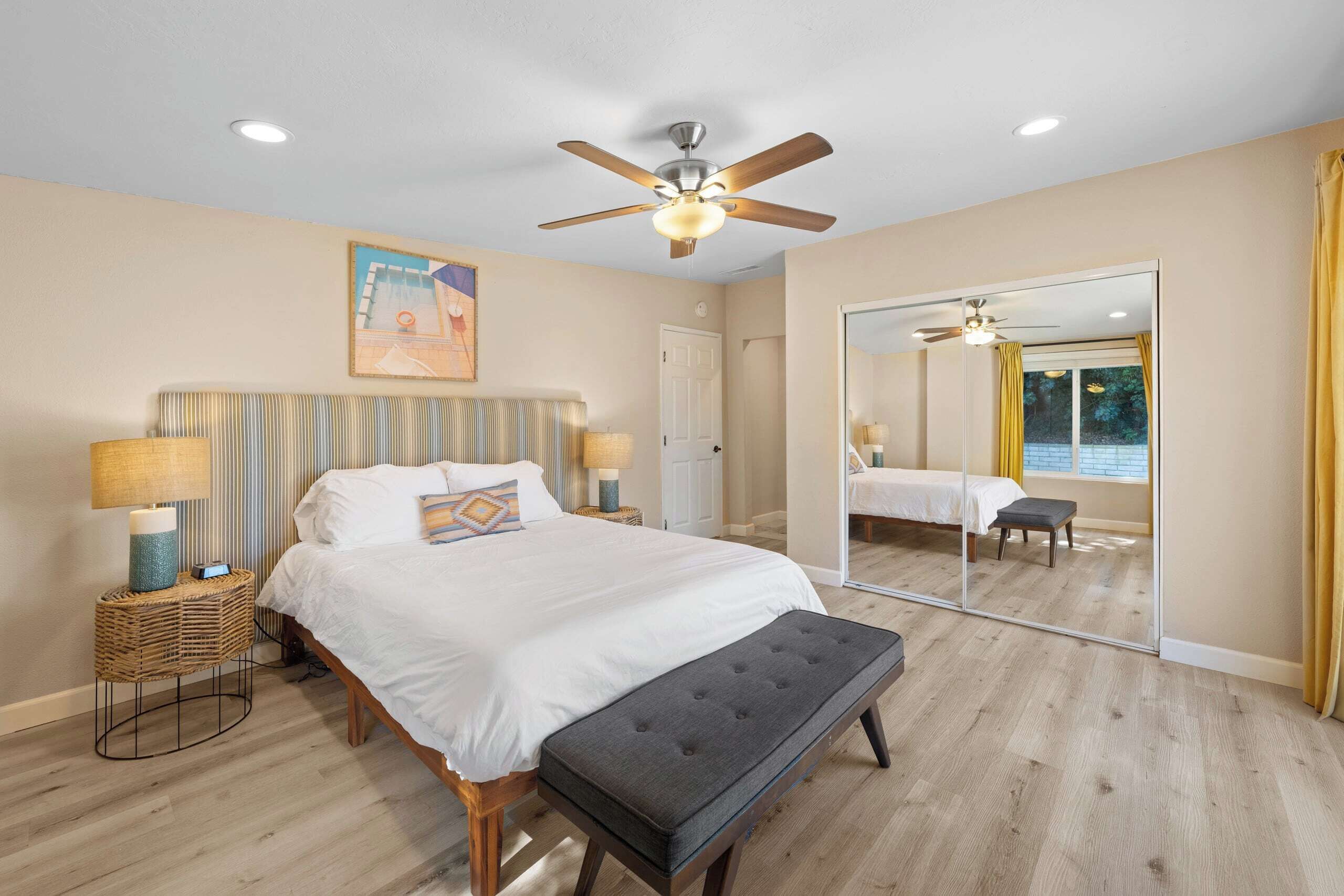 A bedroom with a bed, bedside lamps, a ceiling fan, a large mirror, and a view of a pool through the window.