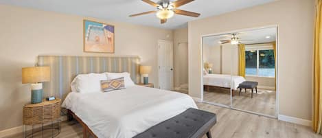 A bedroom with a bed, bedside lamps, a ceiling fan, a large mirror, and a view of a pool through the window.