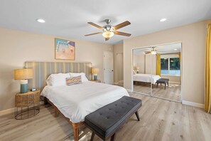 Ein Schlafzimmer mit einem Bett, Nachttischlampen, einem Deckenventilator, einem großen Spiegel und Blick auf einen Pool durch das Fenster.