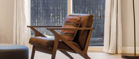 Una habitación con piso de madera, una silla marrón con cojín, un pequeño otoman, una lámpara de pie y vistas a través de una ventana con cortinas.