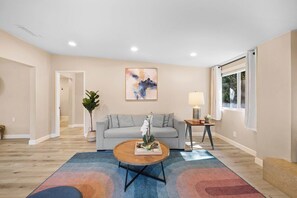 A living room with a grey sofa, a coffee table, a painting on the wall, and a window with curtains. - Spacious and Serene North Park Home (San Diego)