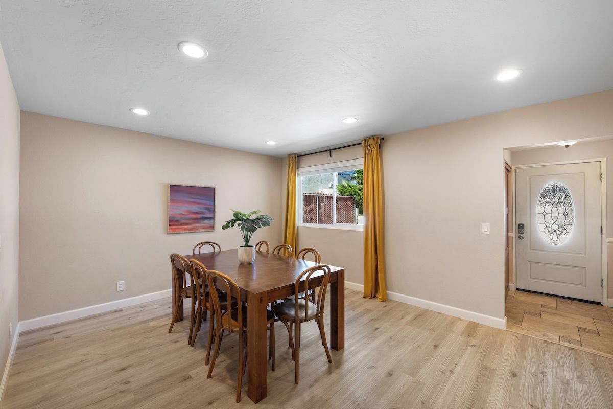 Comedor con mesa y sillas de madera, una planta en maceta y una ventana con cortinas amarillas.