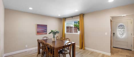 A dining room with a wooden table and chairs, a potted plant, and a window with yellow curtains.