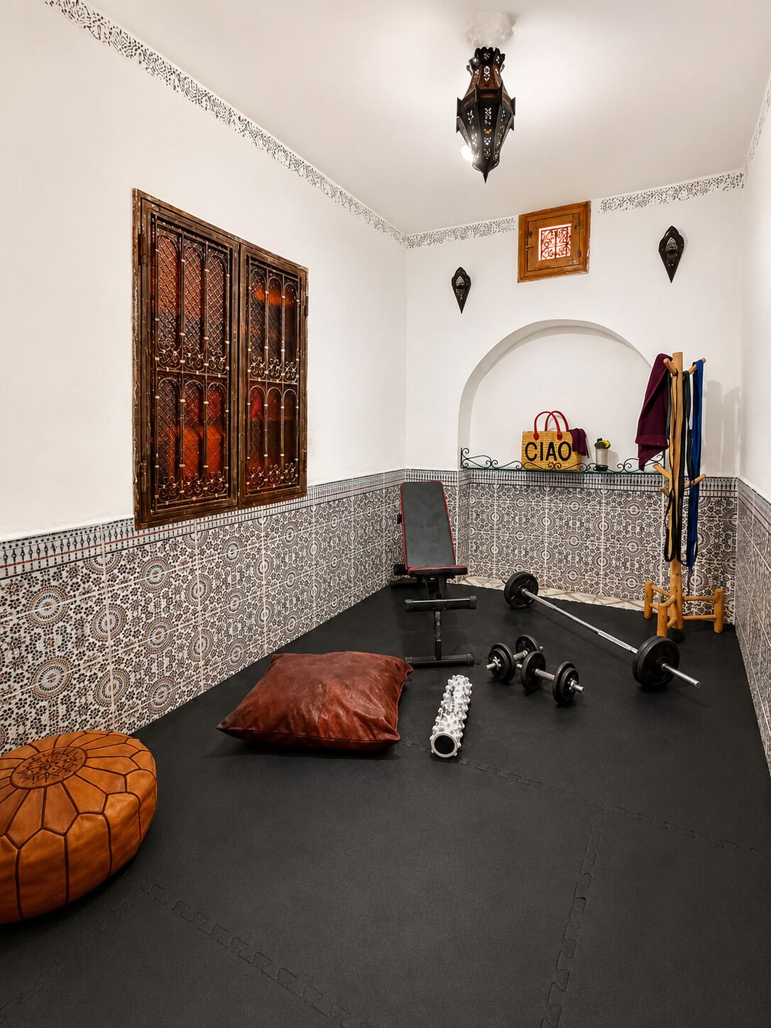 A gym with a black rubber floor, weightlifting equipment, a bench, and a decorative wall with a window and patterned tiles.