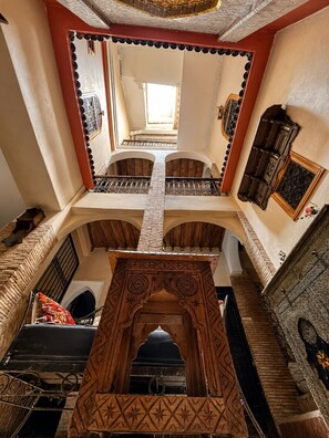 Un interior de varios niveles con balcones arqueados, un arco de madera central y una vista a través de una ventana hacia un espacio exterior.