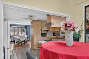 Un comedor con mantel rojo, un jarrón con flores y una cocina al fondo.