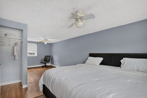 A bedroom with a bed, a ceiling fan, a chair, a window, and a wall-mounted shelf with towels.
