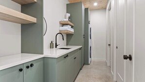 A modern kitchen with a white countertop, a sink, and a built-in oven. There are cabinets with black handles and a wooden shelf above the sink. - Slo | Luxury, Spa & 2 Saunas Memphremagog lake (Stanstead)