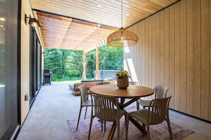 A modern interior with a round wooden table, chairs, a hanging light fixture, and a view of a forested area. - Slo | Luxury, Spa & 2 Saunas Memphremagog lake (Stanstead)