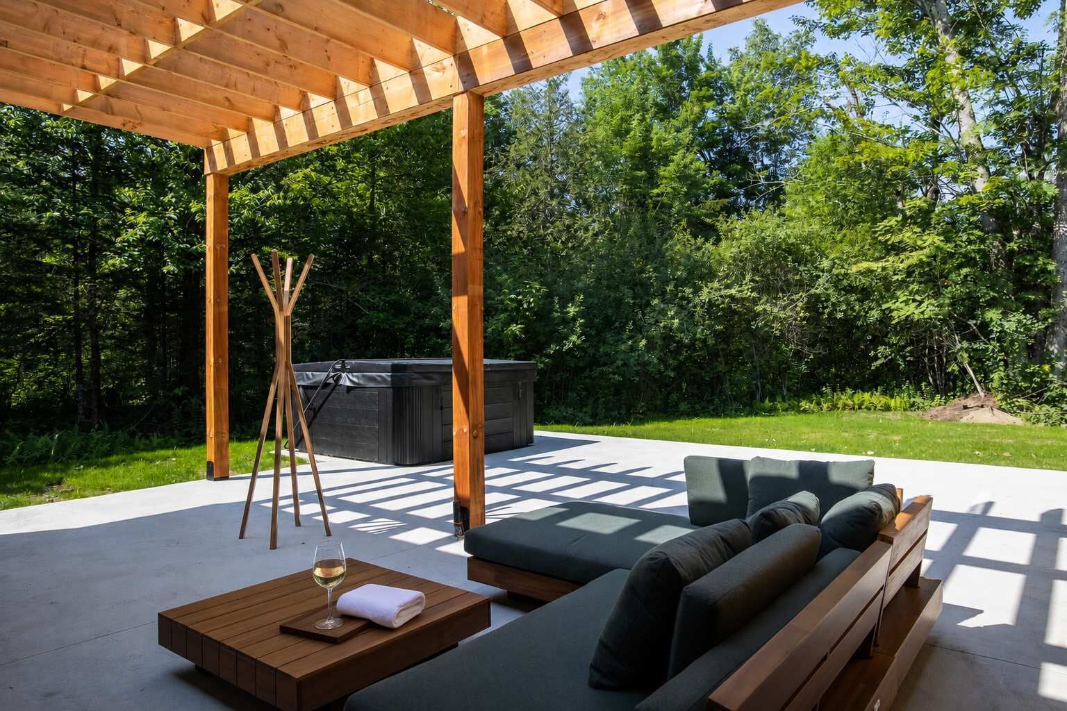Um pátio externo com pérgola de madeira, área de estar com almofadas, mesa com uma taça de vinho e uma banheira de hidromassagem ao fundo.