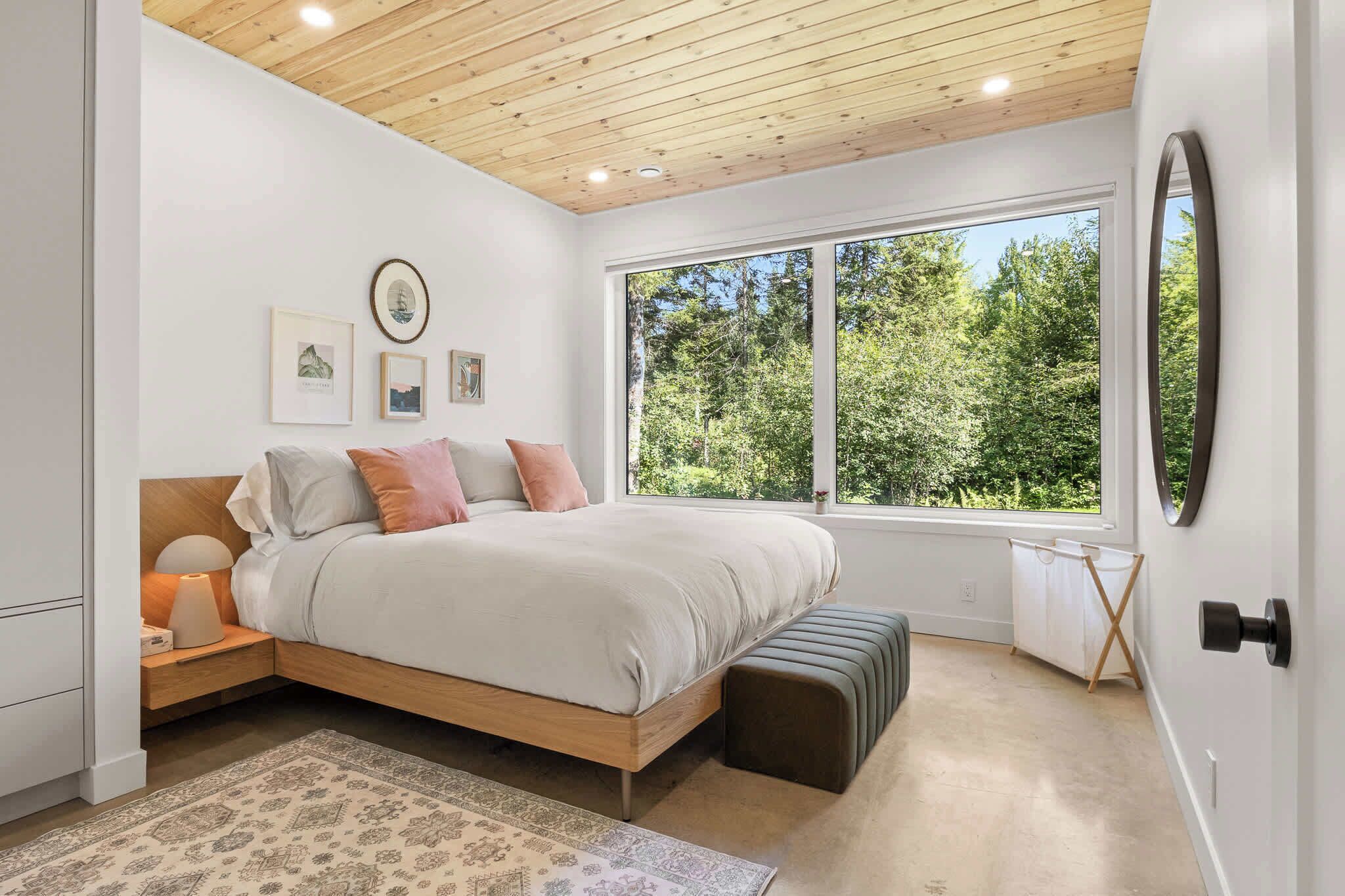 Une chambre avec un grand lit, une table de chevet, une commode, une fenêtre donnant sur des arbres et un plafond avec des poutres en bois.