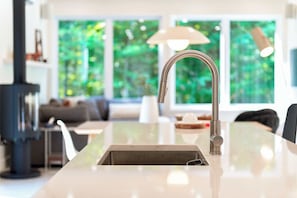 A modern kitchen with a stainless steel faucet, a built-in sink, and a large window offering a view of greenery. - Slo | Luxury, Spa & 2 Saunas Memphremagog lake (Stanstead)
