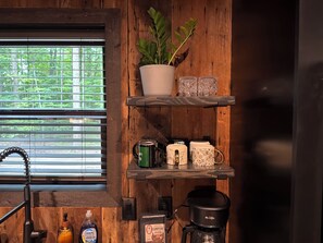 Interior rústico de madeira com uma janela, prateleiras com canecas e uma cafeteira, e uma planta.
