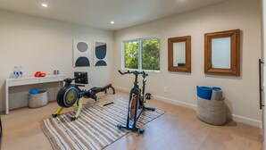Ein Heimfitnessraum mit Trainingsgeräten, ein Schreibtisch mit Wasserflaschen und zwei Fenster mit Blick auf Bäume.