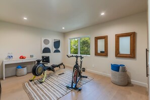 Ein Heimfitnessraum mit Trainingsgeräten, ein Schreibtisch mit Wasserflaschen und zwei Fenster mit Blick auf Bäume.