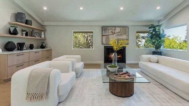 Ein modernes Wohnzimmer mit Kamin, weißen Sofas, einem Glastisch und einem großen Fenster mit Blick auf Bäume.
