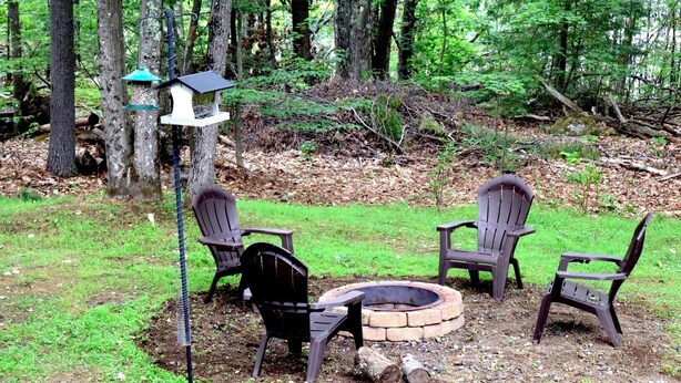 A forested area with a fire pit, chairs, and a bird feeder. - Cozy cabin w/modern amenities in charming Gilford. Pet & family friendly. (Gilford)