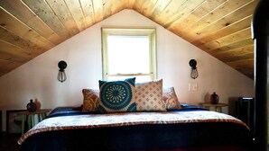 A bedroom with a sloped ceiling, a bed with patterned bedding, two wall-mounted lamps, a window, and a small table with a vase. - Cozy cabin w/modern amenities in charming Gilford. Pet & family friendly. (Gilford)