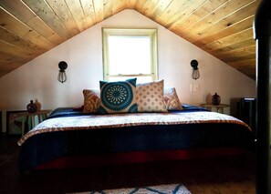 Ein Schlafzimmer mit schrägverdeckter Decke, einem Bett mit gemusterten Betttextilien, zwei Wandleuchten, einem Fenster und einem kleinen Tisch mit einer Vase.