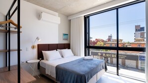 Ein modernes Hotelzimmer mit großem Fenster, einem Bett mit weißer Bettwäsche, einem Nachttisch und einem hängenden Holzgestell.