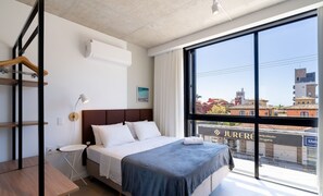 Ein modernes Hotelzimmer mit großem Fenster, einem Bett mit weißer Bettwäsche, einem Nachttisch und einem hängenden Holzgestell.